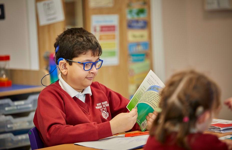 A male pupil in Primary school is sitting at a desk reading a book.
