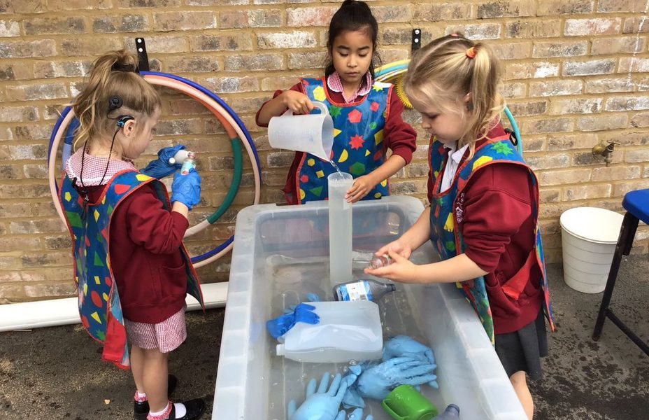 Children are standing at a plastic trough full of water and playing with tubes, containers and rubber gloves