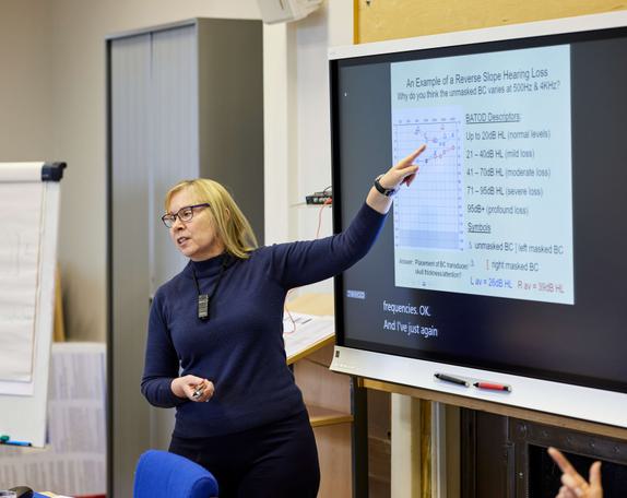 A female lecturer is teaching whilst pointing towards a smart board.