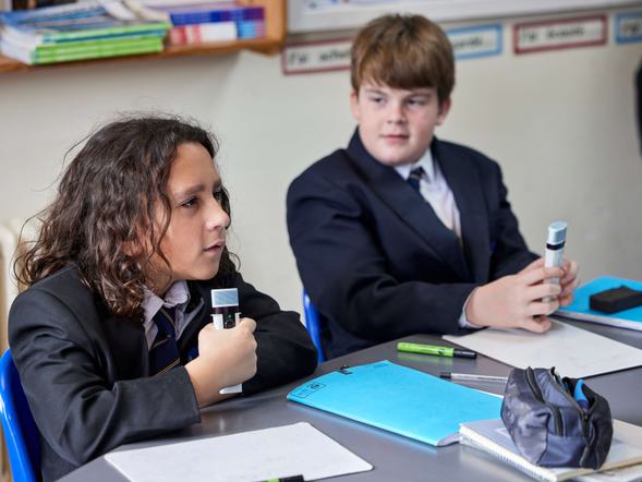 Two young male students are sat in a classroom at their desks. They are holding microphones, one is speaking on the microphone.