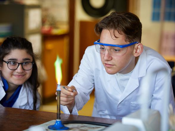 A student is holding some chemicals to a bunsen burner. Another student is watching