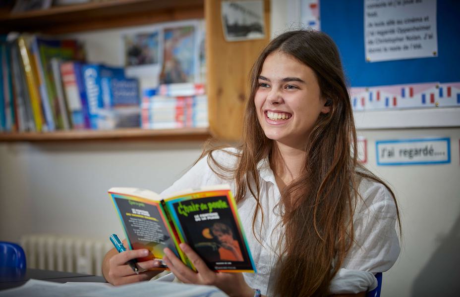 A young female is smiling to someone off camera. She is holding a French fiction book.