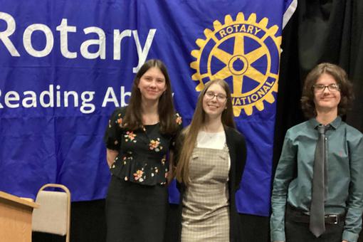 Three students are standing in front of a Rotary flag.