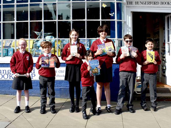 A row of primary pupils are standing in front of a bookshop holding their books and smiling.
