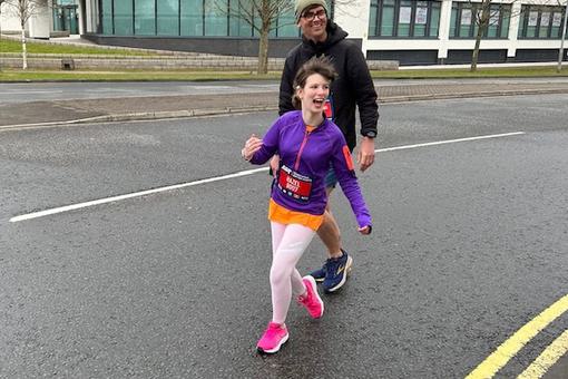 A young female is running with a huge smile on her face. Behind her is a parent.