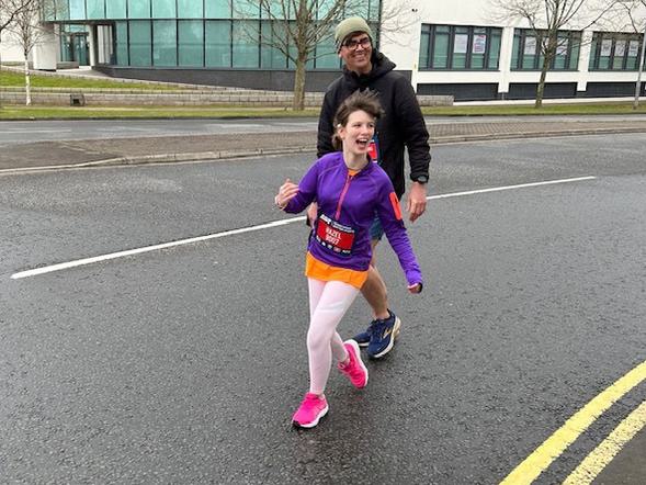 A young female is running with a huge smile on her face. Behind her is a parent.