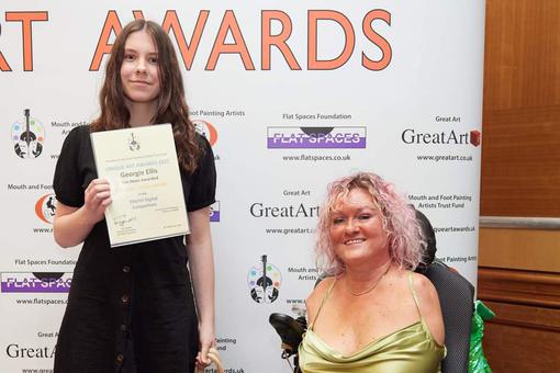 A young female is standing holding a certificate. Next to her an adult female in a wheelchair.