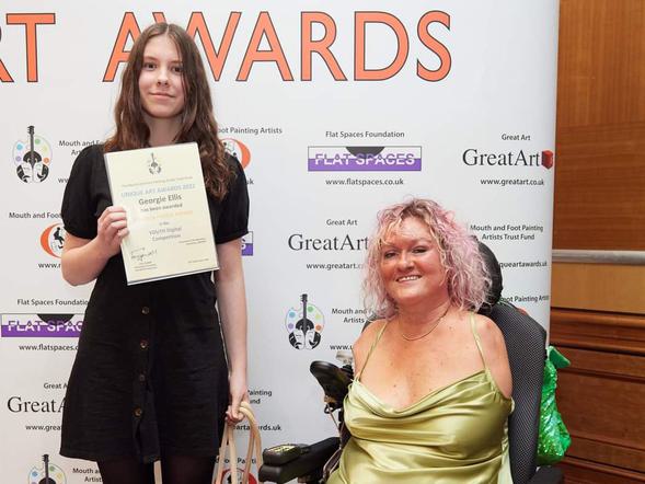 A young female is standing holding a certificate. Next to her an adult female in a wheelchair.